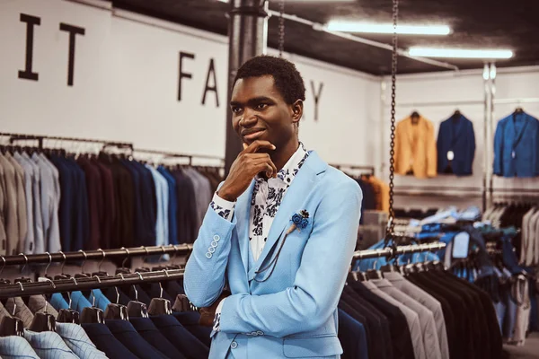 Hombre africano elegante vestido posando con la mano en la barbilla mientras está de pie en una tienda de ropa masculina clásica . — Foto de Stock