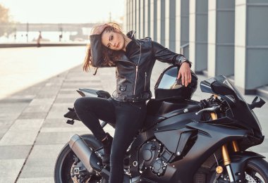 Güzel bir motorcu kız bir binanın dışında süper motoruna yaslanıyor..