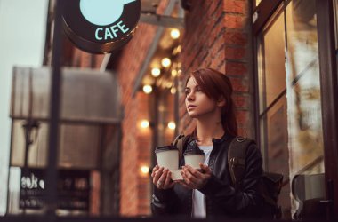 Kafe yakınındaki dışında paket servisi olan restoran kahve bardağı tutan bir sırt çantası ile deri ceketli bir çekici kız.