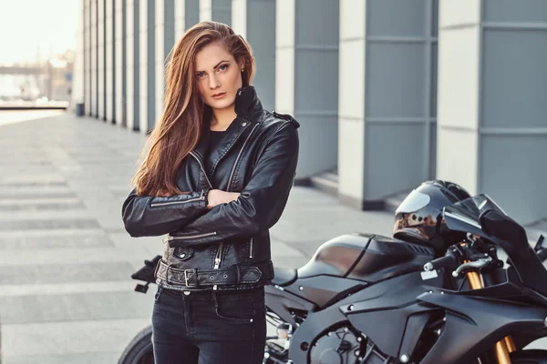 Uma menina motociclista confiante com os braços cruzados ao lado de sua superbike fora de um edifício . — Fotografia de Stock