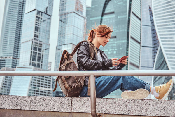 Trendy dressed redhead student girl with tattoos on her face using a smartphone in front of skyscrapers in Moskow city.