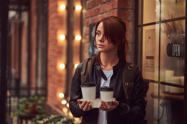 Очаровательная девушка в кожаной куртке с рюкзаком с чашкой кофе на вынос возле кафе . — стоковое фото