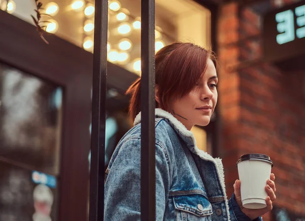 Uma menina bonita vestindo um casaco de ganga segurando xícara com café takeaway fora perto do café . — Fotografia de Stock