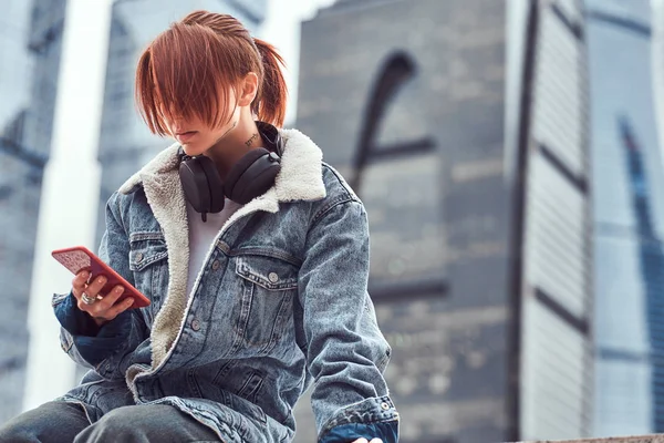 Una chica pelirroja hipster con tatuaje en la cara usando un teléfono inteligente sentado frente a rascacielos en la ciudad de Moskow en la mañana nublada . —  Fotos de Stock