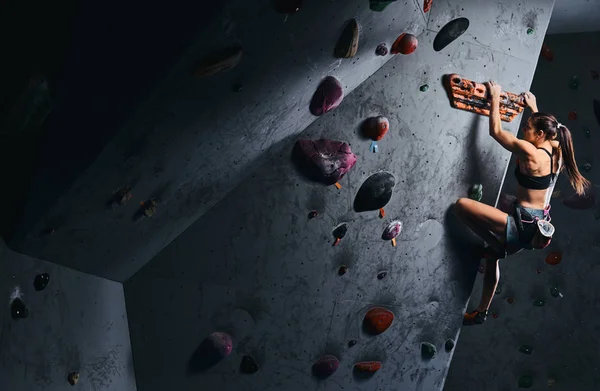 Mujer joven en pantalones cortos y sujetador deportivo haciendo ejercicio en una pared de bouldering en el interior . — Foto de Stock