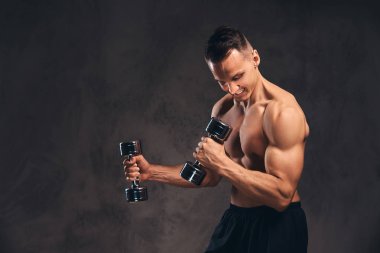Koyu arka plan üzerinde dumbbells ile egzersiz yaparak kas vücut ile genç gömleksiz vücut geliştirmeci.