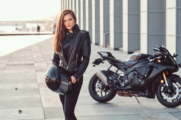 Krásná biker dívka drží helmu vedle superbike mimo budovu. — Stock fotografie