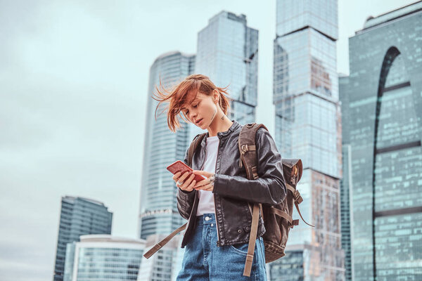 Trendy dressed redhead student girl with tattoos on her face using a smartphone in front of skyscrapers in Moskow city.