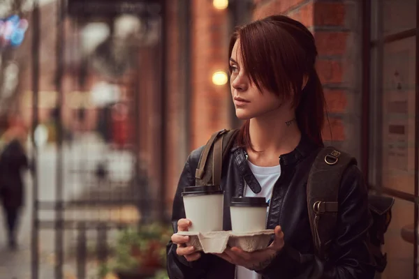 Uma menina encantadora vestindo uma jaqueta de couro com uma mochila segurando xícara com café takeaway fora perto do café . — Fotografia de Stock