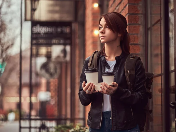 Okouzlující dívka, která nosí koženou bundu s batohem drží pohár s s sebou kávu mimo poblíž kavárny. — Stock fotografie