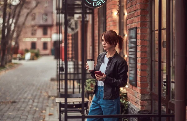 Красивая девушка в кожаной куртке с кофе на вынос и смартфоном возле кафе . — стоковое фото