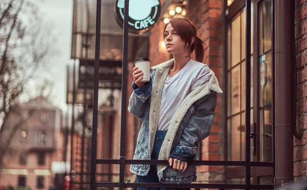 Uma menina bonita vestindo um casaco de ganga segurando xícara com café takeaway fora perto do café . — Fotografia de Stock