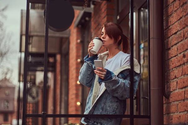 デニム コートを着て美しい刺青の女の子がカフェの外のスマート フォンを持ってテイクアウト コーヒーを飲む. — ストック写真