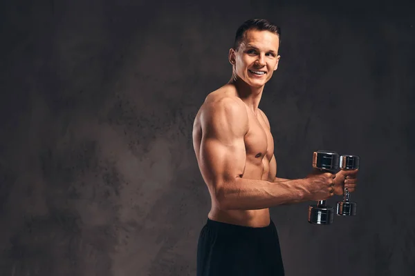 Joven culturista sin camisa con cuerpo musculoso haciendo ejercicio con pesas sobre fondo oscuro . — Foto de Stock