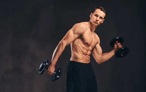 Jeune bodybuilder torse nu avec corps musclé faisant de l'exercice avec haltères sur fond sombre . — Photo