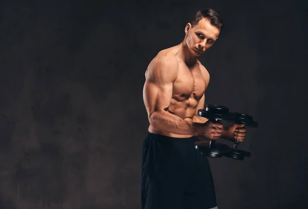 Joven culturista sin camisa con cuerpo musculoso haciendo ejercicio con pesas sobre fondo oscuro . — Foto de Stock