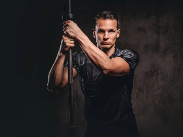 Portret van een knappe jonge bodybuilder holding een barbell en camera kijken op donkere achtergrond. — Stockfoto