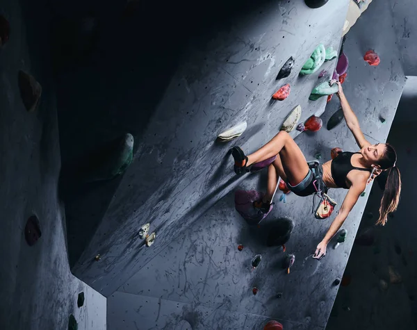 Young woman in shorts and sports bra exercising on a bouldering wall indoors. — Stock Photo, Image