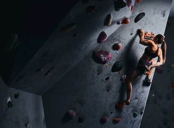 Mujer joven en pantalones cortos y sujetador deportivo haciendo ejercicio en una pared de bouldering en el interior . — Foto de Stock