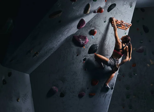 Junge Frau in kurzen Hosen und Sport-BH beim Training an einer Boulderwand drinnen. — Stockfoto