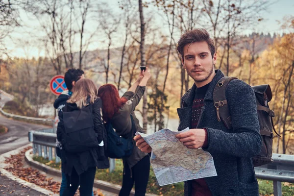 Skupina mladých turistů, stojící na okraji silnice na krásné podzimní les, chlap s mapou plánování výlet. — Stock fotografie