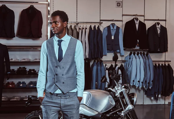 Elegant gekleideter afrikanisch-amerikanischer junger Mann posiert in der Nähe von Retro-Sportmotorrädern im Herrenbekleidungsgeschäft. — Stockfoto