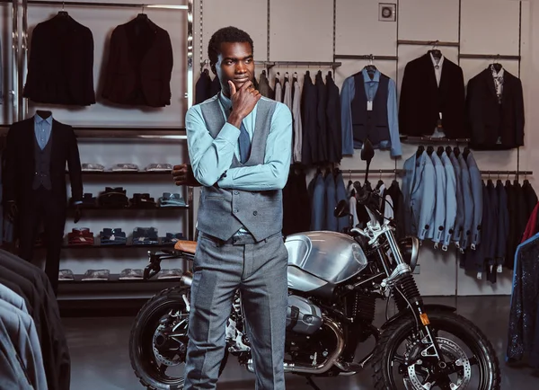 Элегантно одетый афроамериканец позирует возле ретро-спортивного мотоцикла в магазине мужской одежды . — стоковое фото