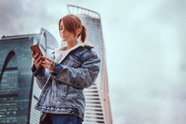 Şık giyimli hippi kız dövmeler yüzü ve bulutlu sabah Moskova şehir içinde gökdelenler önünde bir smartphone kullanarak el ile.