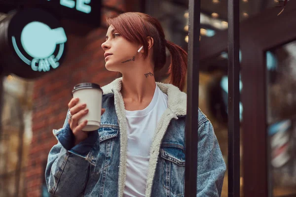 A beautiful girl wearing a denim coat holding cup with takeaway coffee outside near the cafe. — Stock Photo, Image