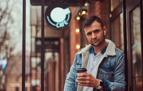 Un hombre guapo y elegante con una chaqueta de mezclilla con auriculares inalámbricos sosteniendo café para llevar fuera de la cafetería . — Foto de Stock