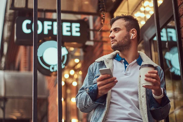 Un hombre guapo y elegante con una chaqueta de mezclilla con auriculares inalámbricos sosteniendo café para llevar fuera de la cafetería . — Foto de Stock