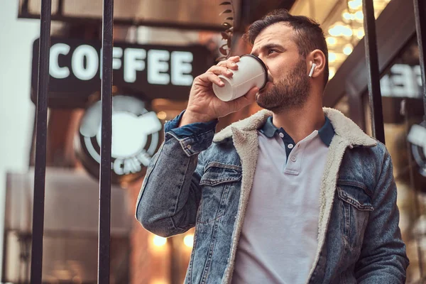 Un hombre guapo y elegante con una chaqueta de mezclilla con auriculares inalámbricos sosteniendo café para llevar fuera de la cafetería . — Foto de Stock
