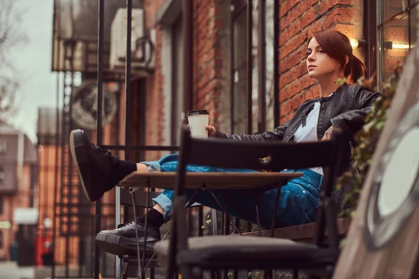 Zrzka dívka, která nosí módní oblečení sedí mimo kavárnu a nápoje káva s sebou. — Stock fotografie