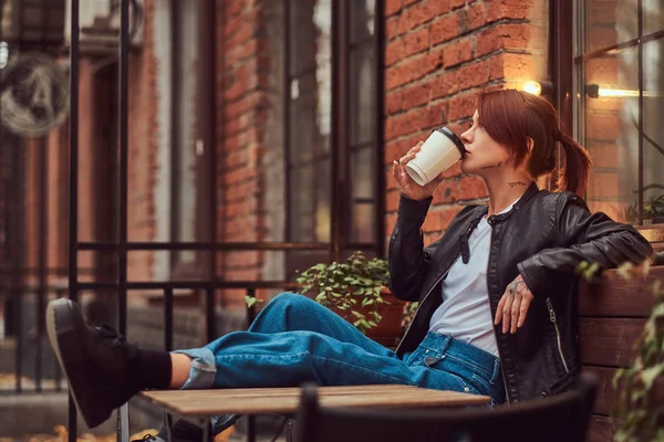 Rödhårig flicka klädd i trendiga kläder sitter utanför café och drycker takeaway kaffe. — Stockfoto