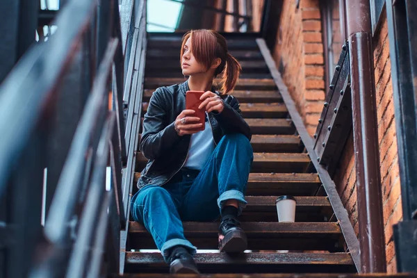 Krásná rusovláska dívka, která nosí módní oblečení sedí na schodech před kavárnou a držení smartphone. — Stock fotografie