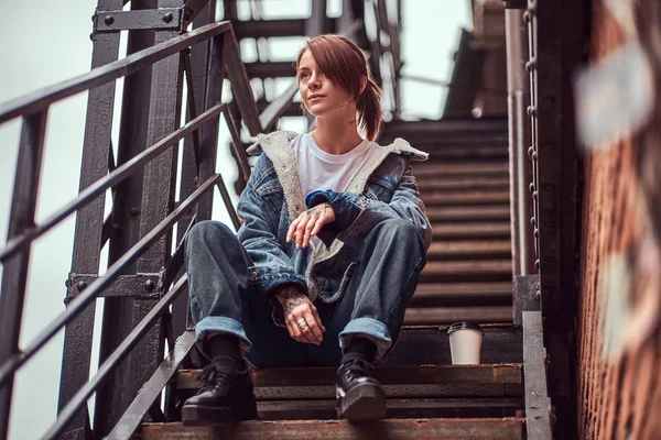 Ein tätowiertes Mädchen in trendiger Kleidung, das draußen auf der Treppe sitzt. — Stockfoto