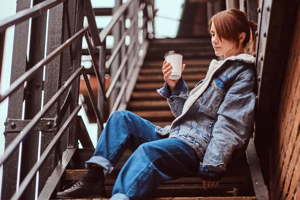 Una hermosa chica con ropa de moda sentada en las escaleras fuera de la cafetería y sosteniendo el café para llevar . —  Fotos de Stock