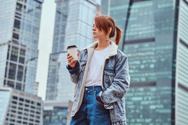 Trendiga klädd hipster tjej med tatuering på hennes ansikte som håller takeaway-kaffe framför skyskrapor i Moskow city. — Stockfoto