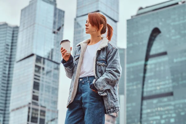 Stijlvolle roodharige hipster meisje met tatoeage op haar gezicht dragen denim jasje afhaalmaaltijden koffie houden voor wolkenkrabbers in Moskow stad. — Stockfoto