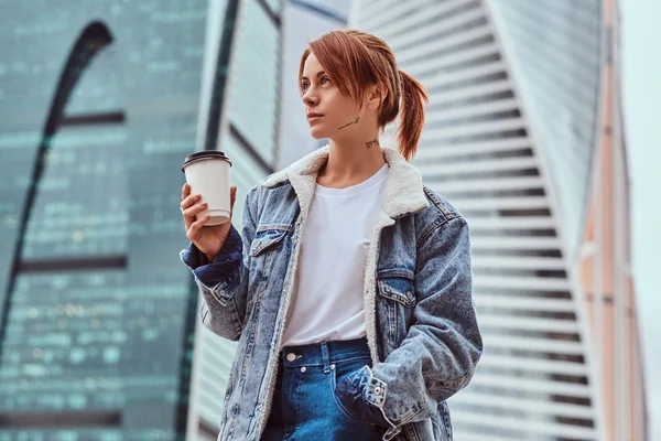 Stylish redhead hipster girl with tattoo on her face wearing denim jacket holding takeaway coffee in front of skyscrapers in Moskow city. — Stock Photo, Image