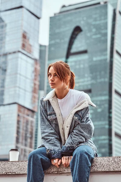 Eleganta rödhårig hipster tjej med tatuering på hennes ansikte bär denim jacka sittande framför skyskrapor i Moskow city på mulen morgon. — Stockfoto