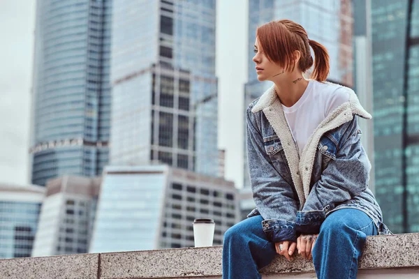 Menina hipster ruiva elegante com tatuagem em seu rosto usando jaqueta de ganga sentado na frente de arranha-céus na cidade de Moskow na manhã nublada . — Fotografia de Stock