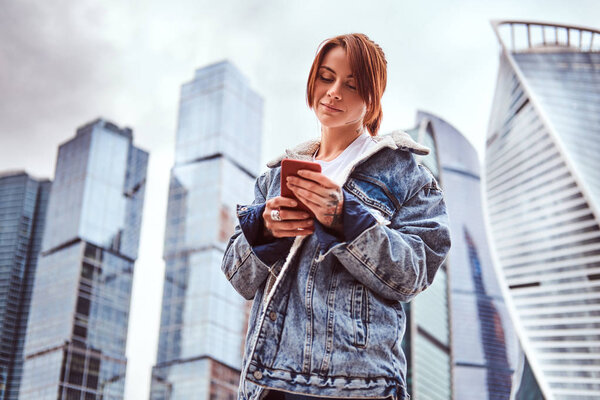 Trendy dressed hipster girl with tattoos on her face and hand using a smartphone in front of skyscrapers in Moskow city at cloudy morning.
