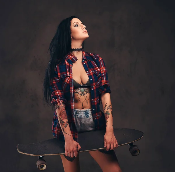 Sexy ragazza tatuata indossa una camicia a scacchi sbottonata rossa e pantaloncini che tengono uno skateboard in posa in uno studio . — Foto Stock