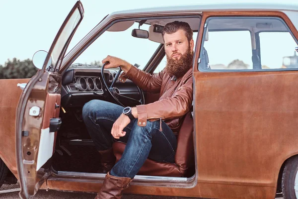 Бородатий чоловік, одягнений у коричневу шкіряну куртку, сидить за кермом налаштованого ретро-автомобіля з відкритими дверима . — стокове фото