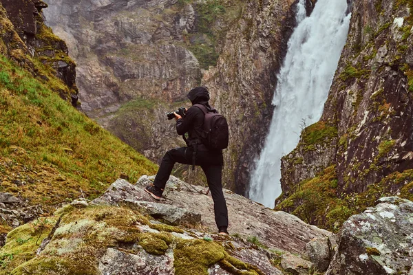 Nature photographer tourist with camera shoots while standing on the mountain against a waterfall. — Stock Photo, Image