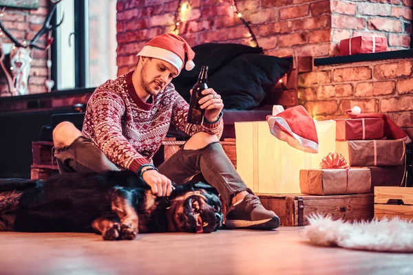 A stylish man holding a bottle with beer while sitting with his cute dog in a decorated living room at Christmas time.