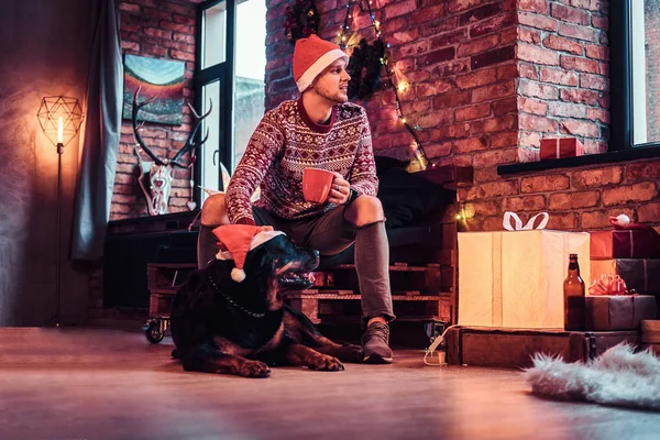 A young man holding cup with coffee during Christmas time with his cute dog in a decorated living room. — Stock Photo, Image