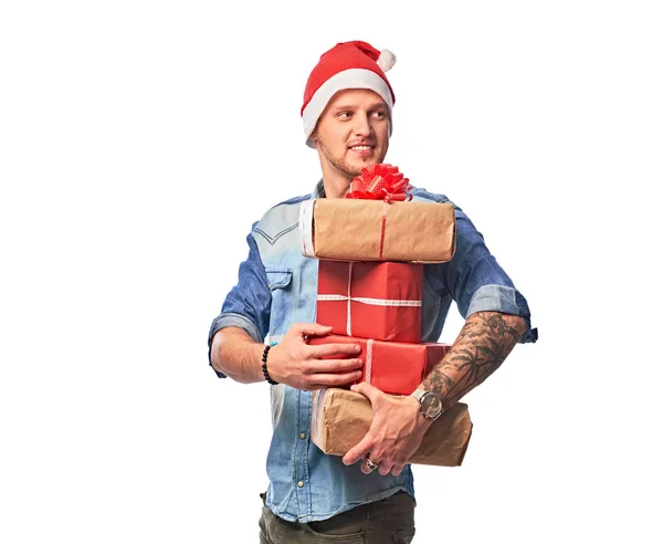 Un hombre feliz vistiendo una camisa de mezclilla y un sombrero de Santa Claus sosteniendo cajas de regalos . — Foto de Stock