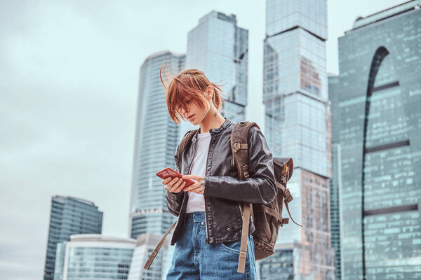 Trendy dressed redhead student girl with tattoos on her face using a smartphone in front of skyscrapers in Moskow city.